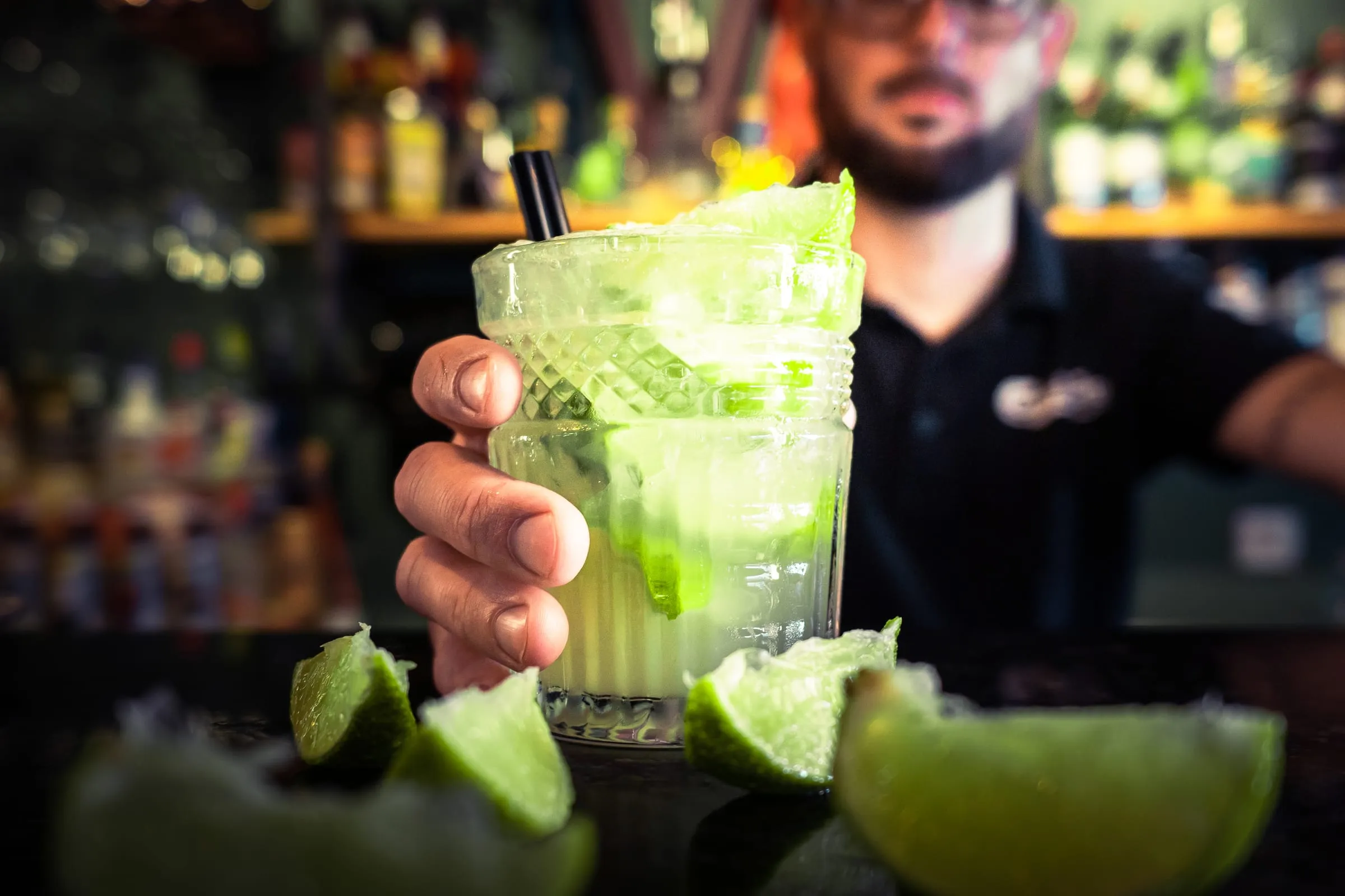 Barman estende um mojito por cima do balcão, com limas em primeiro plano.